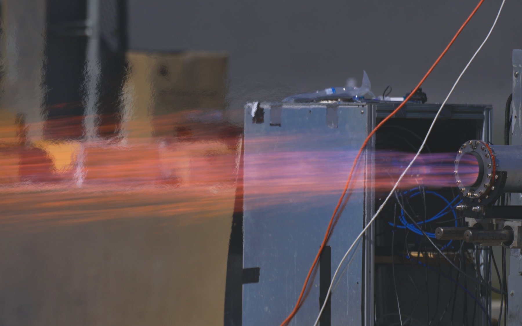 The engine at full thrust on the horizontal test stand, a long orange plume with a blue flame at the nozzle extending to the left