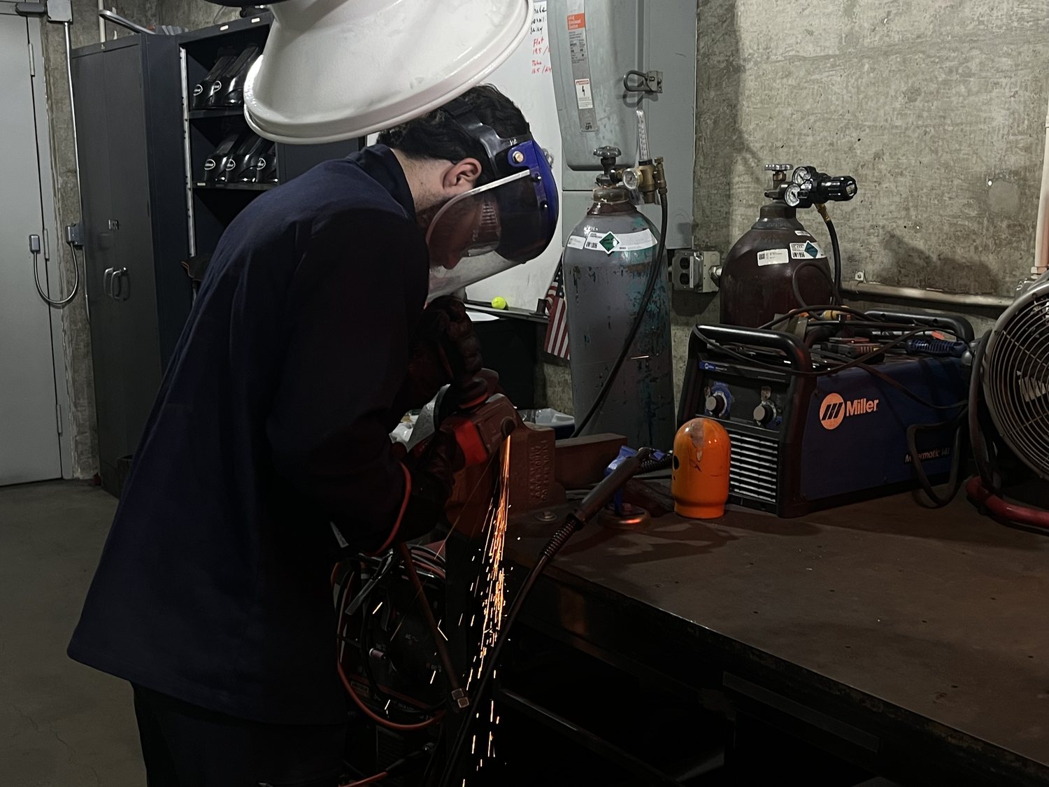 Aras in a hard hat and face shield grinding a steel part, sparks flying, at a bench in the machine shop