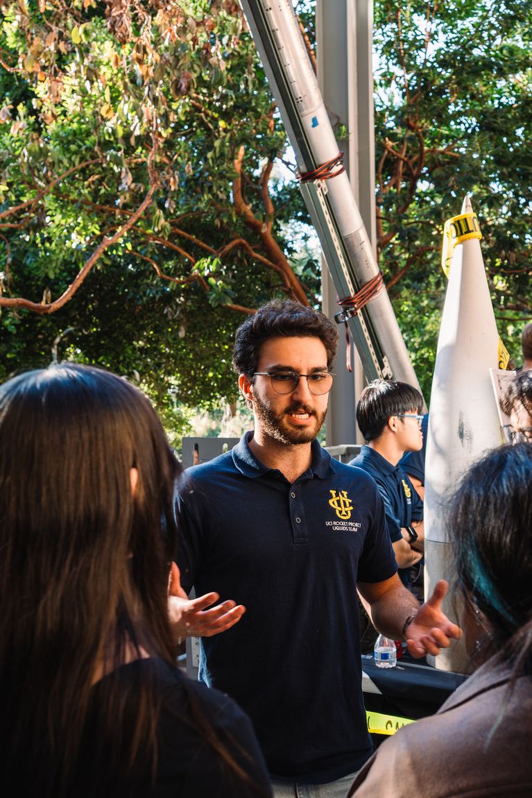 Aras in a UCI Rocket Project polo talking with prospective members at an outdoor recruitment table, a rocket airframe and a nose cone on display behind him